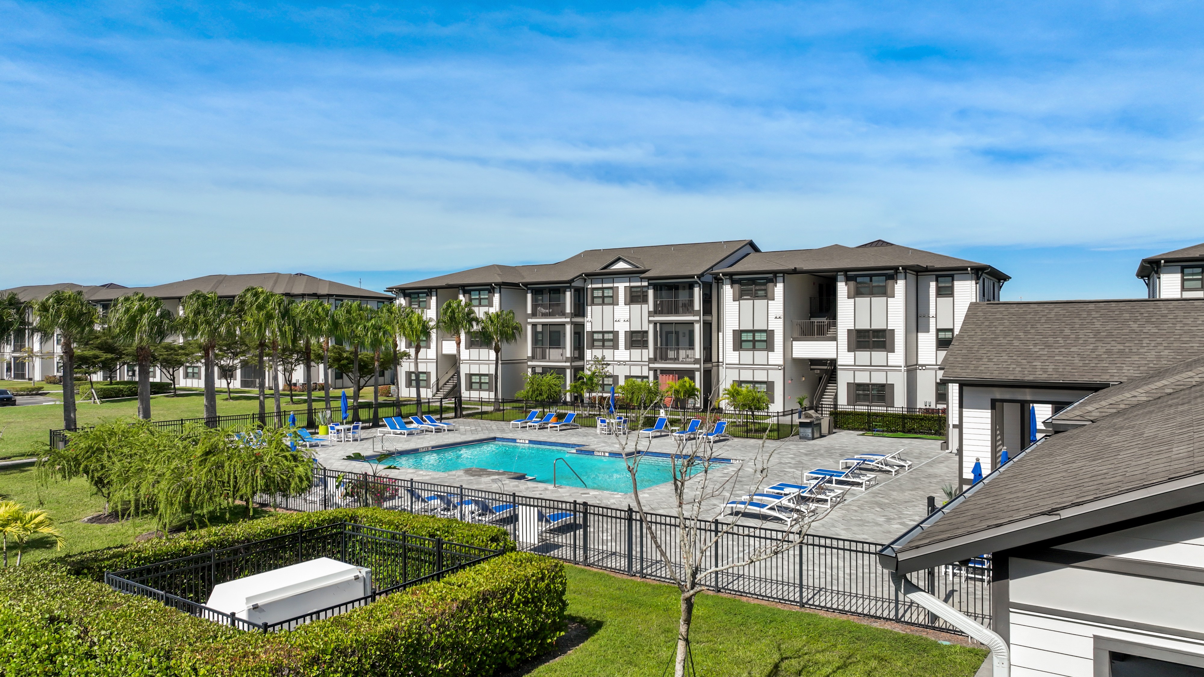 A large swimming pool surrounded by lounge chairs and a fence with a building in the background.